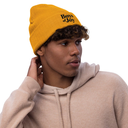Model wearing mustard ribbed beanie with cuff and ‘Boys of Joy’ logo, casual pose in beige hoodie.