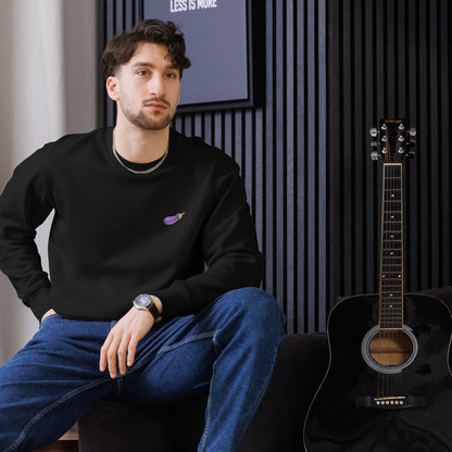 Model seated indoors wearing a black eco sweatshirt with a small purple eggplant graphic on the left chest; acoustic guitar beside him.