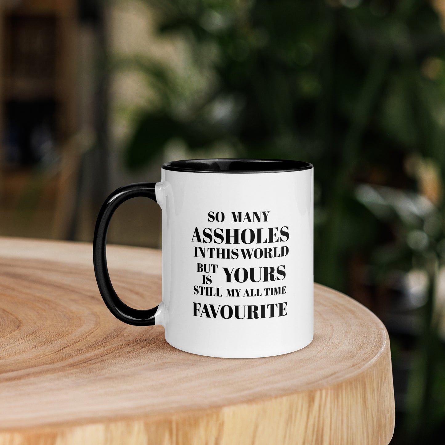 White ceramic mug with black handle and black interior, featuring the quote “SO MANY ASSHOLES IN THIS WORLD BUT YOURS IS STILL MY ALL TIME FAVOURITE,” on a wooden table with a blurred green background.