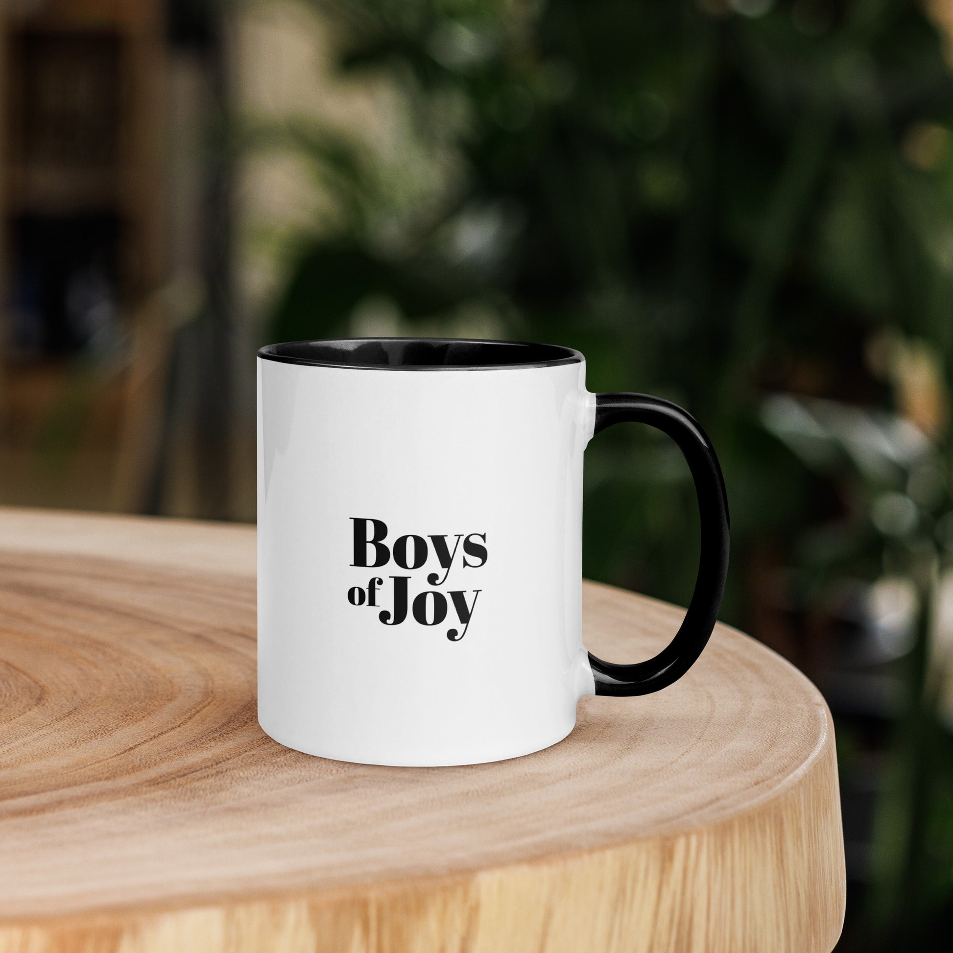 White ceramic mug with black handle and black interior, “Boys of Joy” logo printed on the front, sitting on a wooden table with a blurred green background.